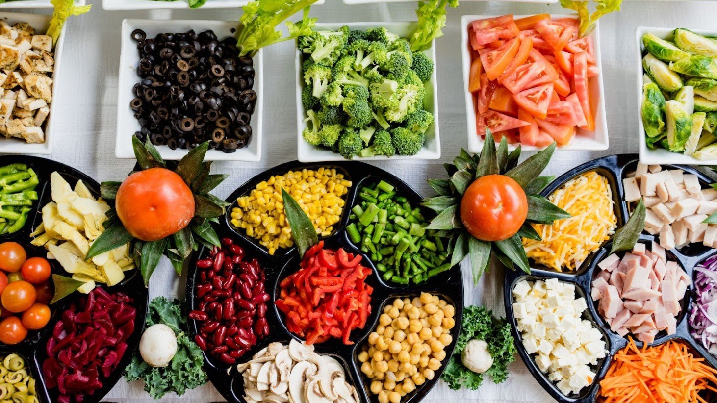 An array of Mediterranean diet ingredients spread out on a table. The center features a tomato with leaves arranged to look like a flower. Surrounding it are separate containers filled with various colorful foods including black olives, diced tomatoes, broccoli florets, green beans, corn kernels, sliced red peppers, kidney beans, chickpeas, shredded cheese, tofu cubes, chopped ham, and sliced mushrooms