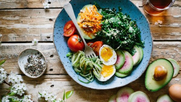 A well-balanced meal displayed on a wooden table consisting of a blue plate with a mix of vibrant salad ingredients including sliced avocados, watermelon radishes, cherry tomatoes, shredded carrots, cucumber noodles, boiled egg halves, and a leafy green salad with sesame seeds sprinkled on top
