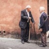 Two elderly people standing next to a lime-washed wall on a cobble road; Researches Unveil the Maximum Age Humans Can Live To